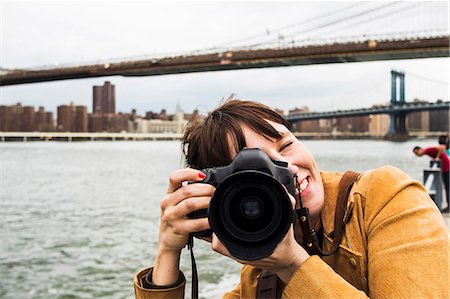 Smiling woman with camera against Brooklyn bridge and East River Stock Photo - Premium Royalty-Free, Code: 6127-08704413