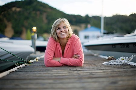 Smiling mid-adult woman lying on her stomach with her arms crossed on a wooden jetty. Photographie de stock - Premium Libres de Droits, Code: 6128-08747537