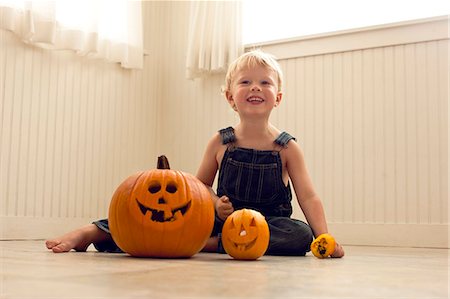 Smiling,  young boy sits a floor behind a big Jack O'Lantern and a small Jack O'Lantern as he poses for a portrait. Fotografie stock - Premium Royalty-Free, Codice: 6128-08747706