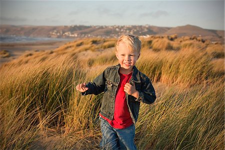 simsearch:6128-08767321,k - Small boy smiles as he standson a grassy sand dune and poses for a portrait. Foto de stock - Royalty Free Premium, Número: 6128-08747710