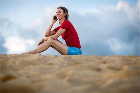 simsearch:6128-08738364,k - Smiling mid adult woman taking on a cell phone while sitting on a sandy beach in wet clothes. Stock Photo - Premium Royalty-Free, Code: 6128-08748129