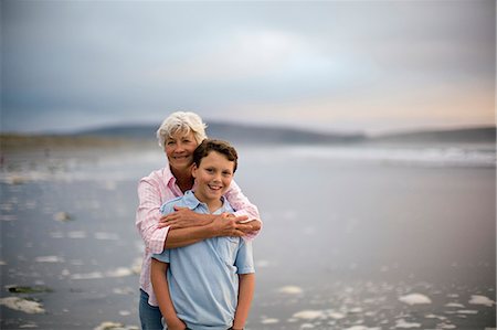 simsearch:6128-08767321,k - Mature woman hugs her grandson from behind as they stand together on a beach and pose for a portrait. Foto de stock - Royalty Free Premium, Número: 6128-08766644