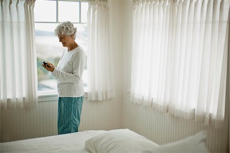 simsearch:614-08031039,k - Mature woman in her pajamas smiles as she looks down at a cell phone as she stands in front of a bedroom window. Stock Photo - Premium Royalty-Free, Code: 6128-08766650