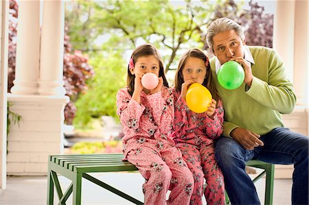 simsearch:6128-08767314,k - Two young girls in matching pajamas sit with a mature man on a bench on a porch and blow up a balloon each as they pose for a portrait. Stock Photo - Premium Royalty-Free, Code: 6128-08766984