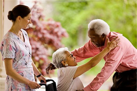 simsearch:6128-08766931,k - Nurse looks on as senior woman in wheelchair reaches up to embrace senior man who is reaching down to hug her. Stock Photo - Premium Royalty-Free, Code: 6128-08766933