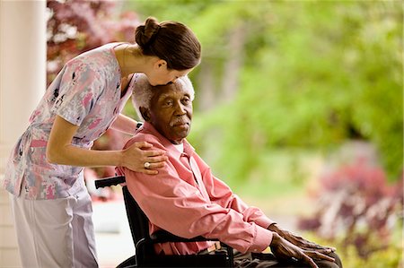 Nurse puts supportive hands on a senior man's shoulders and leans forward to kiss his forehead as he sits in a wheelchair on a porch. Stock Photo - Premium Royalty-Free, Code: 6128-08766934