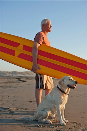 Thoughtful senior man standing on a beach with a surfboard and his dog. Foto de stock - Sin royalties Premium, Código: 6128-08767206