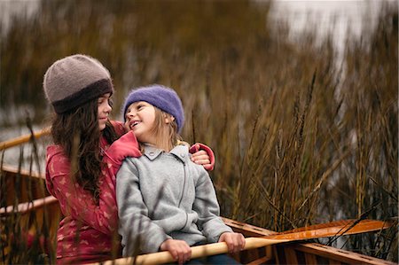 simsearch:6128-08767321,k - Young sisters smile at each other as they sit together in a wooden canoe among reeds on a lakeshore and pose for a portrait. Foto de stock - Royalty Free Premium, Número: 6128-08767318