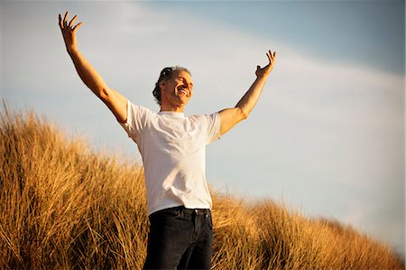 Happy mature man standing on a grassy sand dune with his arms raised. Stock Photo - Premium Royalty-Free, Code: 6128-08780994