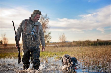 Duck hunter walking through a shallow lake alongside his dog who is holding a dead duck in it's mouth. Photographie de stock - Premium Libres de Droits, Code: 6128-08780995