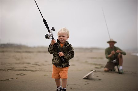 simsearch:700-06758132,k - Smiling young toddler carrying a fishing rod while walking along a beach being watched by his father. Foto de stock - Royalty Free Premium, Número: 6128-08798742
