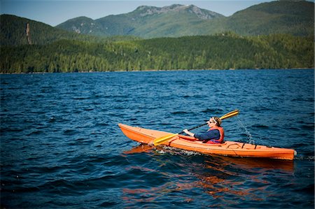 Young woman kayaking with tree covered mountains in the background Stock Photo - Premium Royalty-Free, Code: 6128-08799062