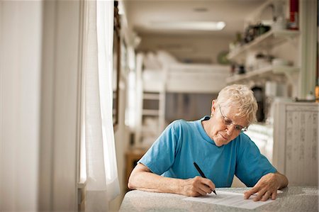 simsearch:6128-08727873,k - Elderly woman signs some paperwork at the kitchen table. Photographie de stock - Premium Libres de Droits, Code: 6128-08727872