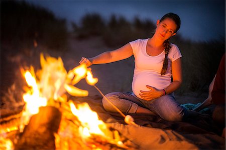 people by camp fire - Pregnant young woman enjoys toasting marshmallows over a beach bonfire at night. Stock Photo - Premium Royalty-Free, Code: 6128-08728013