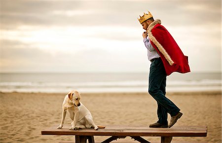 Mid-adult man wearing robe and crown while standing on table at the beach with his dog. Stock Photo - Premium Royalty-Free, Code: 6128-08737583