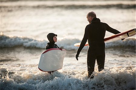 Mid adult man and his young son having a conversation while standing in shallow water holding their boards at the beach. Stock Photo - Premium Royalty-Free, Code: 6128-08738250