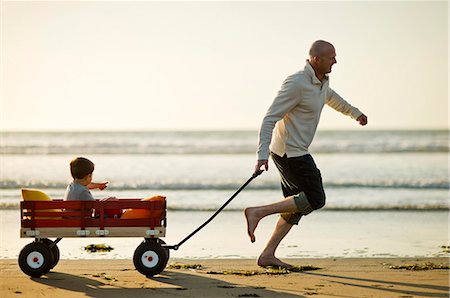 Father pulling his young son along the beach in a little red wagon. Stock Photo - Premium Royalty-Free, Code: 6128-08738550