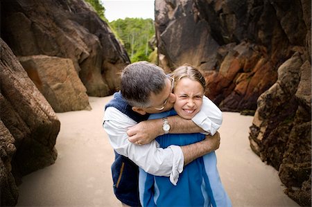 simsearch:6128-08825403,k - Young girl makes a silly face as her father hugs her from behind and kisses her on the cheek as they stand in a sandy cove. Stock Photo - Premium Royalty-Free, Code: 6128-08825384