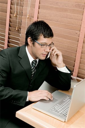 High angle view of a businessman sitting in front of a laptop using a laptop and talking on a mobile phone Stock Photo - Premium Royalty-Free, Code: 625-00849719