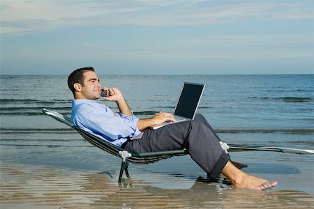 Side profile of a young man sitting on a lounge chair on the beach talking on a mobile phone and using a laptop Stock Photo - Premium Royalty-Free, Code: 625-01094006