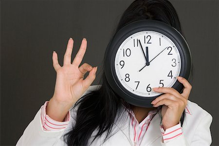 Close-up of a businesswoman holding a clock in front of her face and making an OK sign Stock Photo - Premium Royalty-Free, Code: 625-01748551