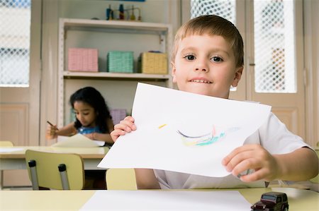 Portrait of a boy showing a drawing with a girl drawing in the background Stock Photo - Premium Royalty-Free, Code: 625-02928944