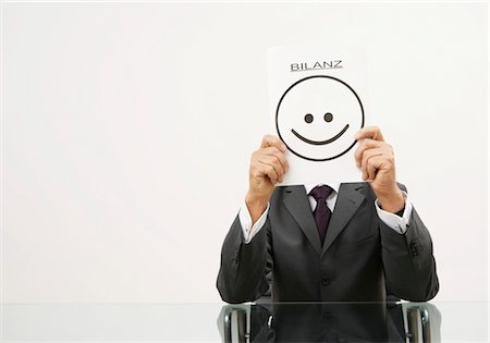 Businessman holding a piece of paper in front of his face with a smiley on it and letters saying "Bilanz" Stock Photo - Premium Royalty-Free, Code: 628-05817414