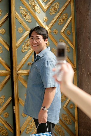 Female hand taking a picture of an Asian man who is standing in front of an ornate door, selective focus Stock Photo - Premium Royalty-Free, Code: 628-00919084