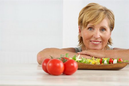 simsearch:628-01495397,k - Femme senior souriant à la caméra, une assiette à salade et tomates au premier plan Photographie de stock - Premium Libres de Droits, Code: 628-01494795