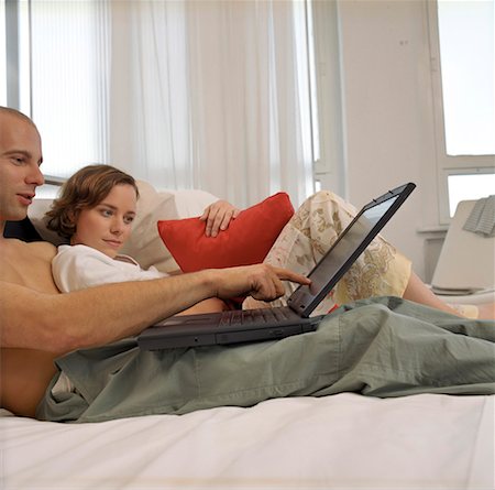 Young woman lying in bed next to a young man, both looking at a laptop Stock Photo - Premium Royalty-Free, Code: 628-01712390