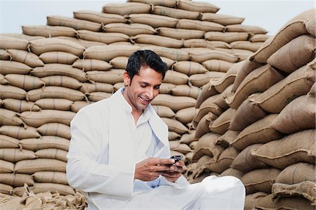 Man sitting on sack of wheat grains reading a SMS on mobile phone, Anaj Mandi, Sohna, Gurgaon, Haryana, India Foto de stock - Sin royalties Premium, Código: 630-07071180