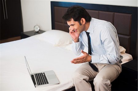 Businessman sitting in front of a laptop on the bed and talking on a mobile phone Stock Photo - Premium Royalty-Free, Code: 630-07071623