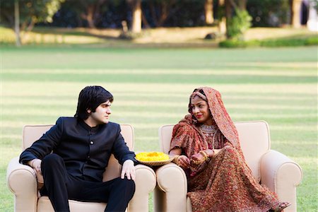 Newlywed couple in traditional wedding dress sitting in armchairs in a lawn and looking at each other Stock Photo - Premium Royalty-Free, Code: 630-01193021