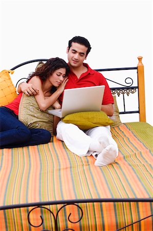 Young man using a laptop on the bed with a young woman leaning on him Stock Photo - Premium Royalty-Free, Code: 630-01297103
