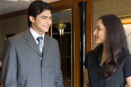 simsearch:630-01708436,k - Businessman and a businesswoman talking to each other and smiling in a hotel Fotografie stock - Premium Royalty-Free, Codice: 630-01708442