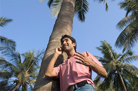 Low angle view of a young man leaning against a tree and talking on a mobile phone Stock Photo - Premium Royalty-Free, Code: 630-01708658