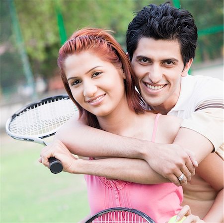 simsearch:625-01037863,k - Portrait d'un jeune homme embrassant une jeune femme par derrière dans un court de tennis et souriant Photographie de stock - Premium Libres de Droits, Code: 630-01874201