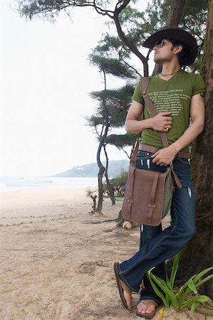 flip flops blue jeans - Young man carrying a shoulder bag and leaning against a tree trunk on the beach Stock Photo - Premium Royalty-Free, Code: 630-01876992