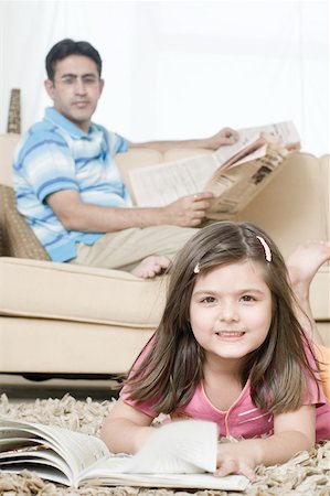 Portrait of a girl lying in front of a book with her father holding newspaper in the background Stock Photo - Premium Royalty-Free, Code: 630-01877498