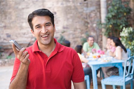 Portrait of a mid adult man holding a mobile phone and smiling with his friends sitting in the background Stock Photo - Premium Royalty-Free, Code: 630-01872829