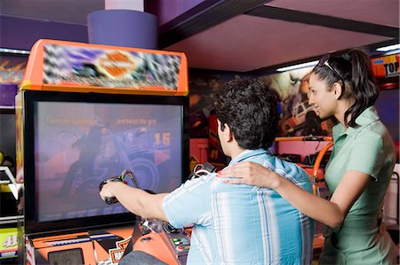 riding a friend on the handlebars - Young man playing video game and a young woman watching his game in a video arcade Stock Photo - Premium Royalty-Free, Code: 630-03481706