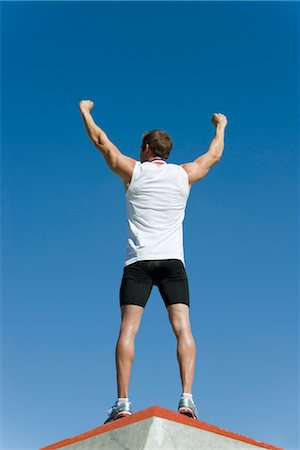 Male athlete standing on winner's podium with arms raised in victory, rear view Stock Photo - Premium Royalty-Free, Code: 632-05759608