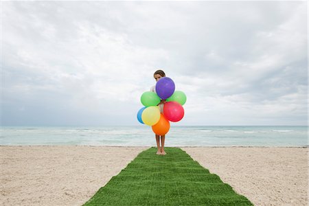 Girl standing on artificial turf on beach, face hiding behind bunch of balloons Stock Photo - Premium Royalty-Free, Code: 632-05759757