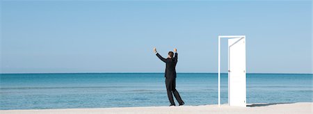 Businessman standing on beach looking at ocean with arms raised, rear view Stock Photo - Premium Royalty-Free, Code: 632-05816432