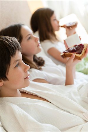 Siblings relaxing on bed, girls eating toasts in background Stock Photo - Premium Royalty-Free, Code: 632-06029881