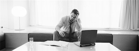 simsearch:695-05772228,k - Businessman sitting holding head, laptop, drink and documents on table in front of him, b&w, panoramic view Stock Photo - Premium Royalty-Free, Code: 632-01143339