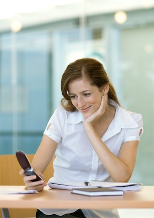 Woman sitting at desk, smiling and looking at cell phone Stock Photo - Premium Royalty-Free, Code: 632-01157612