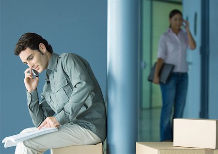 entering office - Young man sitting on cardboard box using cell phone, young woman entering in background Stock Photo - Premium Royalty-Free, Code: 632-01157693