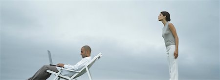 Man sitting in lounge chair with laptop on lap, woman standing peering over man's shoulder, side view Stock Photo - Premium Royalty-Free, Code: 632-01151388