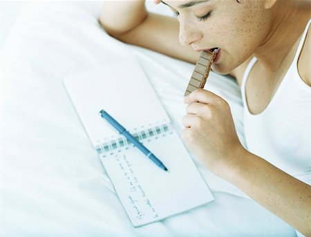 Woman lying on side on bed eating chocolate and looking down at notebook with pen lying across it Stock Photo - Premium Royalty-Free, Code: 632-01151454